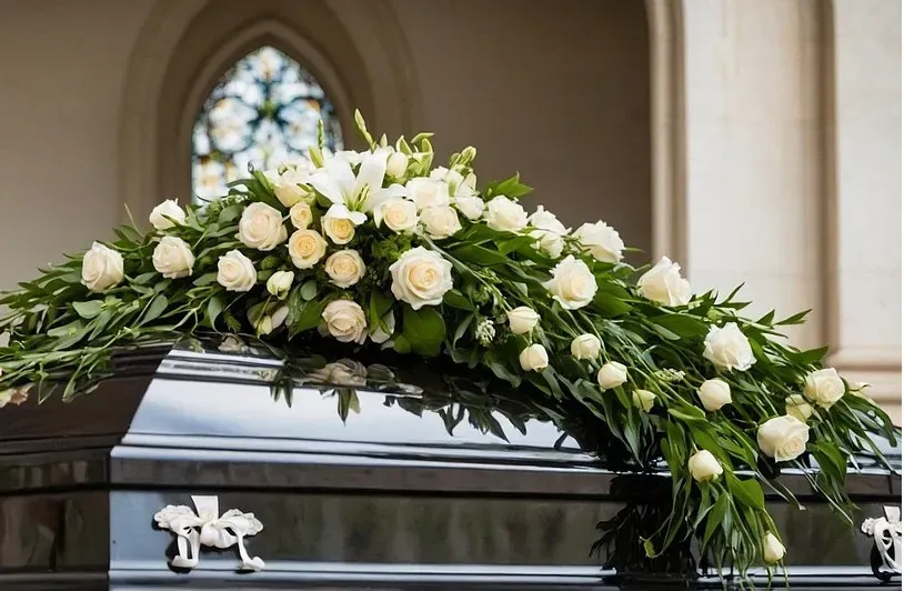 Grand dessus de cercueil composé de roses et lys blancs posé sur un cercueil en bois verni