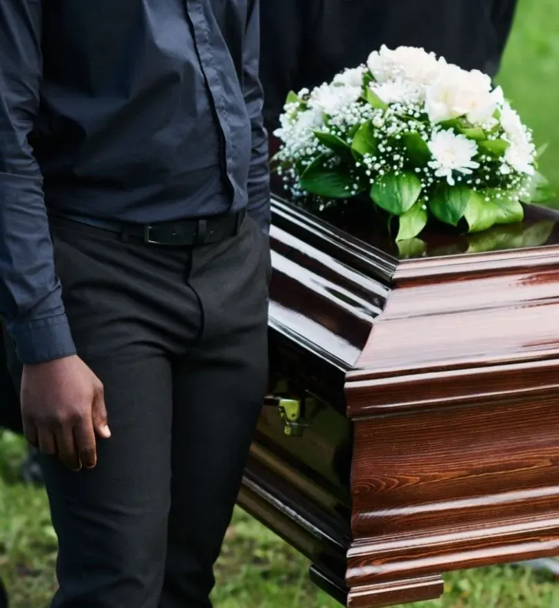Homme en tenue noire debout à côté d'un cercueil en bois verni orné d'un bouquet de fleurs blanches